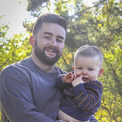 Dr. Collin McCulloch holding a small child close in an outdoor environment with trees and greenery behind them.